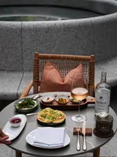 A high-quality, aesthetically pleasing shot of a lunch setting on a terrace at Six Senses London. The image features a well-arranged table with flatbread, dips, and a glass of wine, set against a clean, modern stone background. The lighting is soft and natural, creating an inviting and premium atmosphere.