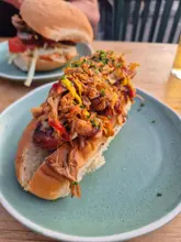 A close-up, high-quality shot of a loaded hot dog topped with pulled pork, crispy onions, and sauces, served on a light blue plate. In the background, slightly out of focus, sits another burger, suggesting a casual meal with a friend. The natural evening lighting enhances the textures of the food, making it look appealing and fresh.