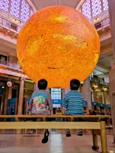 A high-quality, symmetrical photograph capturing two children sitting on a bench in a museum, gazing up at a large, illuminated sculpture of the Sun. The image effectively uses the installation's light to create a silhouette effect, emphasizing the scale of the art and the sense of wonder, while maintaining a clean and professional composition.