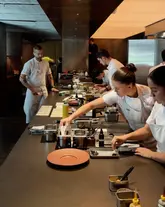 A high-quality, professional shot of the kitchen pass at Ikoyi, capturing the focused and precise nature of the chefs at work. The composition highlights the organized, high-end environment of a Michelin-starred establishment.