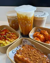 A bright, high-angle flat-lay photograph showcasing a variety of items from a bubble tea and street food venue, including three bubble tea drinks, a portion of Korean-style chicken, dirty waffle fries, and Biscoff French toast.