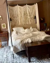 A high-quality, atmospheric shot of a bedroom at Hope Lodge, featuring a rustic four-poster bed with neutral linens and textured wall coverings. The image captures a serene, high-end, and remote aesthetic suitable for a luxury travel destination.