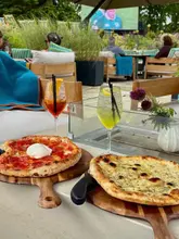 A high-quality, inviting shot of two pizzas and two cocktails on an outdoor terrace, with a sports screen visible in the background, suggesting a relaxed, social atmosphere.