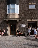A candid, eye-level exterior shot of Toad Bakery in Deptford, capturing the brick facade and the busy atmosphere with customers sitting on benches and entering the shop.