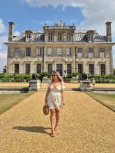 A bright, sunny travel-style photograph featuring a creator standing in front of a grand, historic manor house, likely a National Trust property. The composition is symmetrical and clear, highlighting the scale of the architecture.