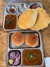 A top-down, flat-lay photograph of two metal trays featuring traditional Indian street food dishes, specifically chole bhature and pav bhaji, set on a wooden table.