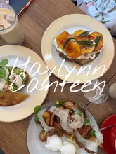A bright, overhead flat-lay shot of a brunch spread at Haylynn Canteen, featuring three distinct dishes and an iced coffee. The composition is clean and inviting, showcasing the vibrant colors of the food against a neutral wooden table.