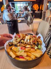 A close-up, eye-level shot of a bowl containing bao buns and fries, served in a casual food hall setting. The background is busy, showing other diners and neon signage, with a cutlery holder prominently placed on the table.
