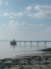 A serene, high-quality landscape photograph of Clevedon Pier stretching out into the calm sea under a soft, blue sky. The image captures the architectural detail of the Victorian pier and the tranquil atmosphere of the coastline, effectively promoting the location as a scenic destination for a relaxing outing.