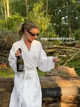 A woman in a white bathrobe sits on a log in a wooded area, holding a bottle of Prosecco and a wine glass with a branded tag, promoting a relaxing countryside retreat experience.