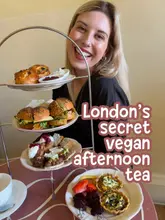 A bright, inviting shot of a creator presenting a vegan afternoon tea spread, effectively combining the venue's food offering with a personal, friendly endorsement.