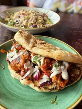 A high-quality, close-up shot of a chicken tikka flatbread served on a green plate, with a chicken and bacon salad visible in the soft-focus background. The natural lighting effectively highlights the textures of the grilled chicken and the fresh herbs.