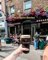 A first-person perspective shot of a pint of Guinness held up in front of 'The Old Stand' pub on a sunny day, capturing a relaxed, social atmosphere.