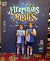A photo of two children posing in front of a branded 'Mermaids & Pirates' theatre backdrop, likely taken by a parent during a family outing to the show.