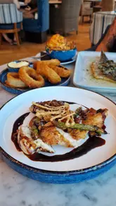 A montage-style reel showcasing a variety of dishes and cocktails at Tir a Môr, a restaurant located within a hotel in Cardiff Bay. The video features close-up shots of food textures, including a cheese pull, breaking a creme brulee, and plating details, ending with a view of the venue's waterfront setting.