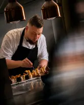 A high-quality, professional photograph capturing a chef meticulously plating intricate dishes in a kitchen environment. The lighting is dramatic and focused, highlighting the precision and artistry involved in the preparation. The image conveys a sense of dedication and high-end culinary skill, typical of a fine dining or private chef service.