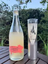 A casual, outdoor shot featuring a bottle of Squealing Pig fuzzy peach wine spritz alongside a Disneyland Paris branded glass, set on a wooden table against a garden background.