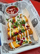 A close-up, top-down shot of a portion of halloumi fries served in a paper tray, topped with a white sauce, pomegranate seeds, and fresh herbs, accompanied by a small pot of red dipping sauce. The food is presented on a newspaper-print liner, which creates a busy visual background.