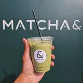 A clean, effective product shot featuring a matcha latte held against a branded green wall. The composition is simple and intentional, placing the product front and center while clearly showcasing the venue's branding in the background.