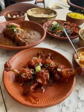A high-quality, well-lit flat lay photograph of an Indian tapas spread at a restaurant. The image showcases a variety of dishes including chicken lollipops, curries, and sides, arranged on a marble table with rustic terracotta plates, effectively communicating the variety and appeal of the menu.