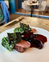 A close-up, eye-level shot of a plated duck breast dish featuring crispy kale and a berry reduction sauce, captured in a restaurant setting.