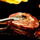 A high-quality, close-up shot of a raw tomahawk steak being held by tongs over a grill with flames in the background. The image is professionally lit and composed, highlighting the texture and quality of the meat, suitable for a culinary tutorial or restaurant promotion.