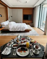 A high-quality, well-composed shot of a luxurious hotel room breakfast spread. The image captures a full breakfast service on a glass table in the foreground, with a neatly made bed and modern, warm-toned interior design in the background. The lighting is soft and natural, creating an inviting and aspirational atmosphere typical of a high-end hospitality experience.