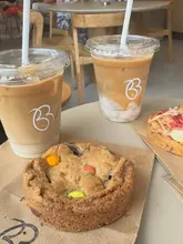 A simple, overhead flat-lay shot featuring two iced lattes and a Smarties-topped cookie on a paper bag, capturing a casual coffee shop visit.