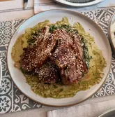 A top-down, overhead shot of a home-cooked meal featuring grilled lamb chops topped with sesame seeds, served over a vibrant green salsa verde. The dish is presented on a ceramic plate set against a patterned placemat, capturing a rustic and appealing dining aesthetic.