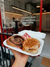 A close-up, first-person perspective shot of a takeaway box containing two artisan buns—a classic cinnamon bun and a Biscoff-topped bun—held in front of the Rise & Bun shopfront in Soho. The image effectively captures the product in a casual, relatable way, highlighting the texture of the buns and the branding on the shop window.