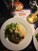 A top-down shot of a meal at a cinema-dining experience, featuring a plate of meatloaf, a bottle of cola, and a bowl of popcorn, alongside a themed menu card.