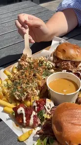 A close-up, top-down shot of a BBQ 'munch box' featuring loaded fries, burgers, and a dipping sauce, captured in an outdoor setting with a hand holding a wooden fork.