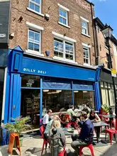 A bright, inviting exterior shot of Dilly Deli in Stockport, capturing the venue's curb appeal with customers enjoying outdoor seating on a sunny day. The image effectively showcases the atmosphere and the welcoming nature of the establishment.