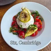 A high-quality, overhead food photography shot of a burrata salad featuring roasted figs and cherry tomatoes. The lighting is soft and natural, highlighting the textures of the cheese and the freshness of the produce. The composition is clean and centered, effectively showcasing the dish against a simple, non-distracting wooden background.