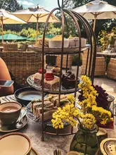 A high-angle shot of an afternoon tea service presented on a birdcage-style stand, set on a table in an outdoor garden. The spread includes sandwiches, cakes, and scones, accompanied by a cup of tea and a vase of yellow flowers.