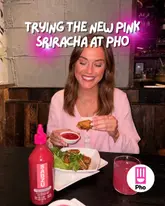 A social media influencer is featured in a restaurant setting, promoting a limited-edition 'Pink Sriracha' sauce at a Pho restaurant. The image shows the creator holding a chicken wing dipped in the sauce, with the bottle of sauce and a pink drink on the table.