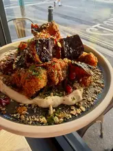 A close-up, appetizing shot of a plate featuring crispy fried aubergine and panko-crumbed buttermilk chicken, served on a base of tahini, garnished with herbs and pomegranate seeds. The photo is taken in a restaurant setting by a window.