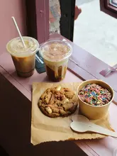 A high-angle, natural-light shot of a coffee shop spread featuring two iced coffees, a large cookie, and a small chocolate dessert pot, arranged on a rustic brown paper surface.