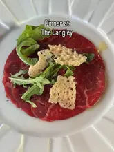 An overhead, centered shot of a beef carpaccio dish served on a white plate, featuring thin slices of raw beef, arugula, and parmesan crisps, with text overlay indicating the location.
