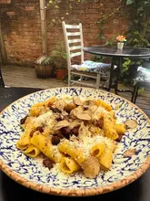 A high-quality, appetizing close-up shot of a truffle carbonara pasta dish served on a patterned plate, set against an outdoor patio background. The image effectively highlights the texture of the pasta and the generous amount of truffle shavings, creating a strong visual appeal for the venue.