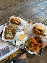 A top-down, POV-style shot of a tray containing two burgers, a portion of chicken tenders, and a portion of loaded chicken, served on a rustic wooden table at a burger restaurant.
