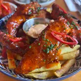 A high-quality, close-up food shot featuring Nashville-style chicken tenders glazed in a vibrant red sauce, served over a portion of fries. The dish is garnished with fresh chives and sliced red chili, presented in a patterned bowl with a side of dipping sauce, effectively showcasing the texture and appetizing nature of the meal.