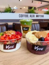 A casual, brightly lit shot of two build-your-own bowls (yogurt and acai) and a matcha drink from a cafe, presented in plastic takeaway containers.