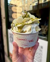 A close-up, handheld shot of a pistachio soft-serve gelato topped with white chocolate buttons, served in a branded paper cup. The image is taken at a 45-degree angle, effectively showcasing the texture and toppings of the dessert against a softly blurred street background.