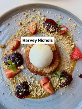 A high-quality, top-down photograph of a plated Pain Perdu dessert served at Harvey Nichols. The dish is elegantly presented with a scoop of ice cream, fresh strawberries, a berry compote, and a dusting of crushed pistachios on a grey plate, set against a clean, neutral background.