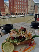 A casual, lifestyle-focused shot of a brunch dish featuring chorizo and eggs on toast, served with coffee, captured at an outdoor table overlooking a harbour.