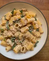 A top-down, well-lit shot of a bowl of rigatoni pasta tossed in a creamy sauce with sausage and spinach, served on a simple wooden surface.