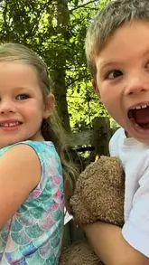 A candid, high-energy selfie of two children holding a teddy bear in an outdoor setting, capturing the joy of a family day out.