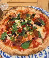 A high-quality, top-down shot of a freshly baked pizza topped with veggie nduja, sliced chillies, and fresh basil, served on a patterned plate on a wooden table with a wine glass in the background.