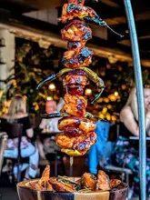 A close-up, eye-level shot of a signature hanging kebab dish served at a restaurant, featuring charred meat and vegetables, with a bowl of potato wedges in the foreground. The background is a blurred, lively restaurant interior with diners and warm lighting.